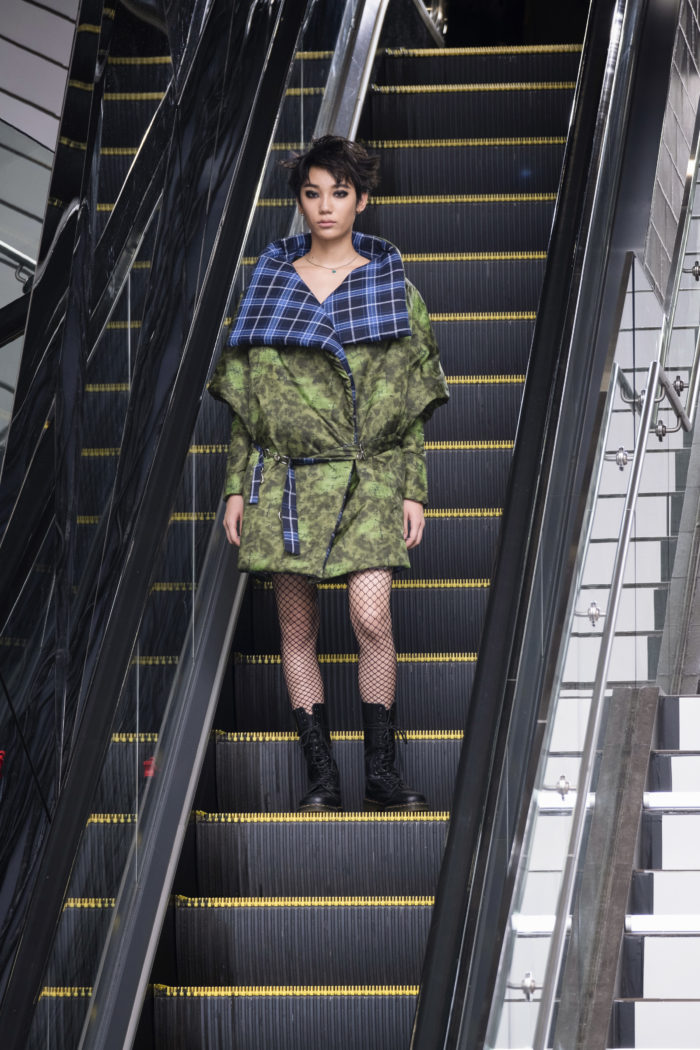 Model on an escalator wearing a green print puffer jacket with contrasting blue-and-black plaid neckline and collar facing