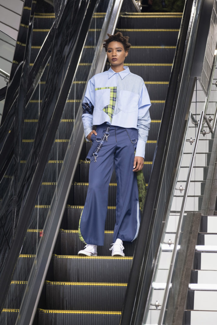 Model on escalator in streetwear, a loose-fitting button-front top and gray pants, and white sneakers