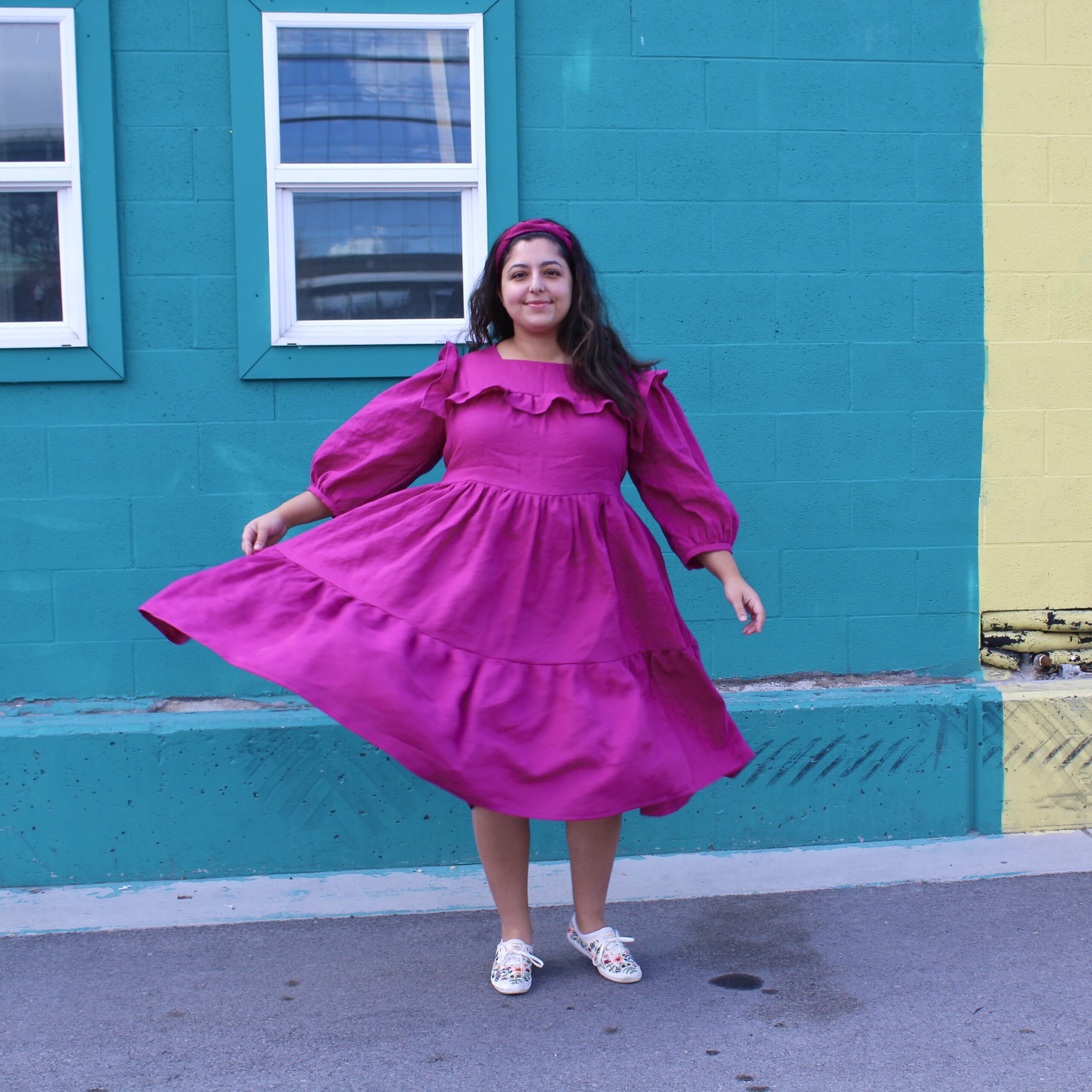 Romy, a POC, mid twirl in her bright magenta ruffle tiered dress smiling in front of a green wall.