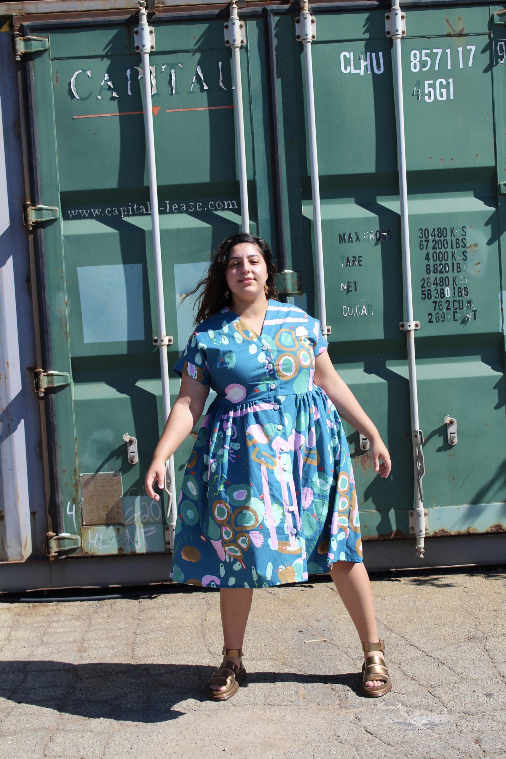 Romy, a POC, wearing a green dress with pink, ochre, lighter green abstract shapes and brass earrings, standing tall in an industrial setting. 