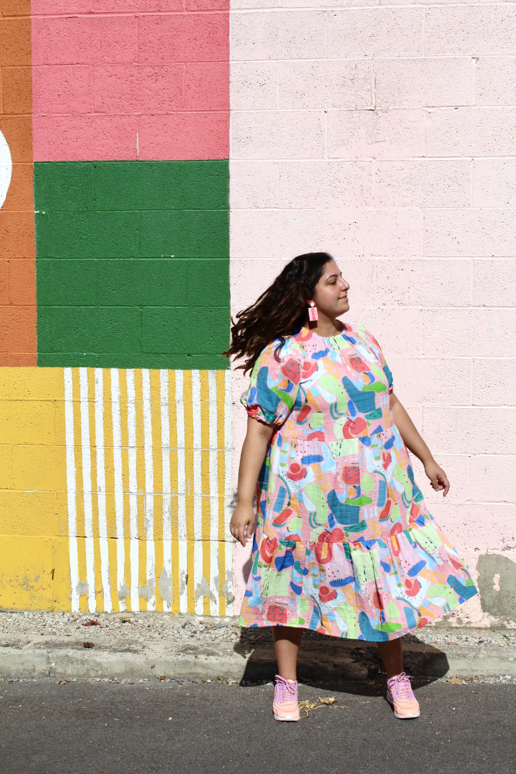 @sewlike, Romy-Krystal Cutler, in a bold printed dress, in front of a colorfully painted wall.