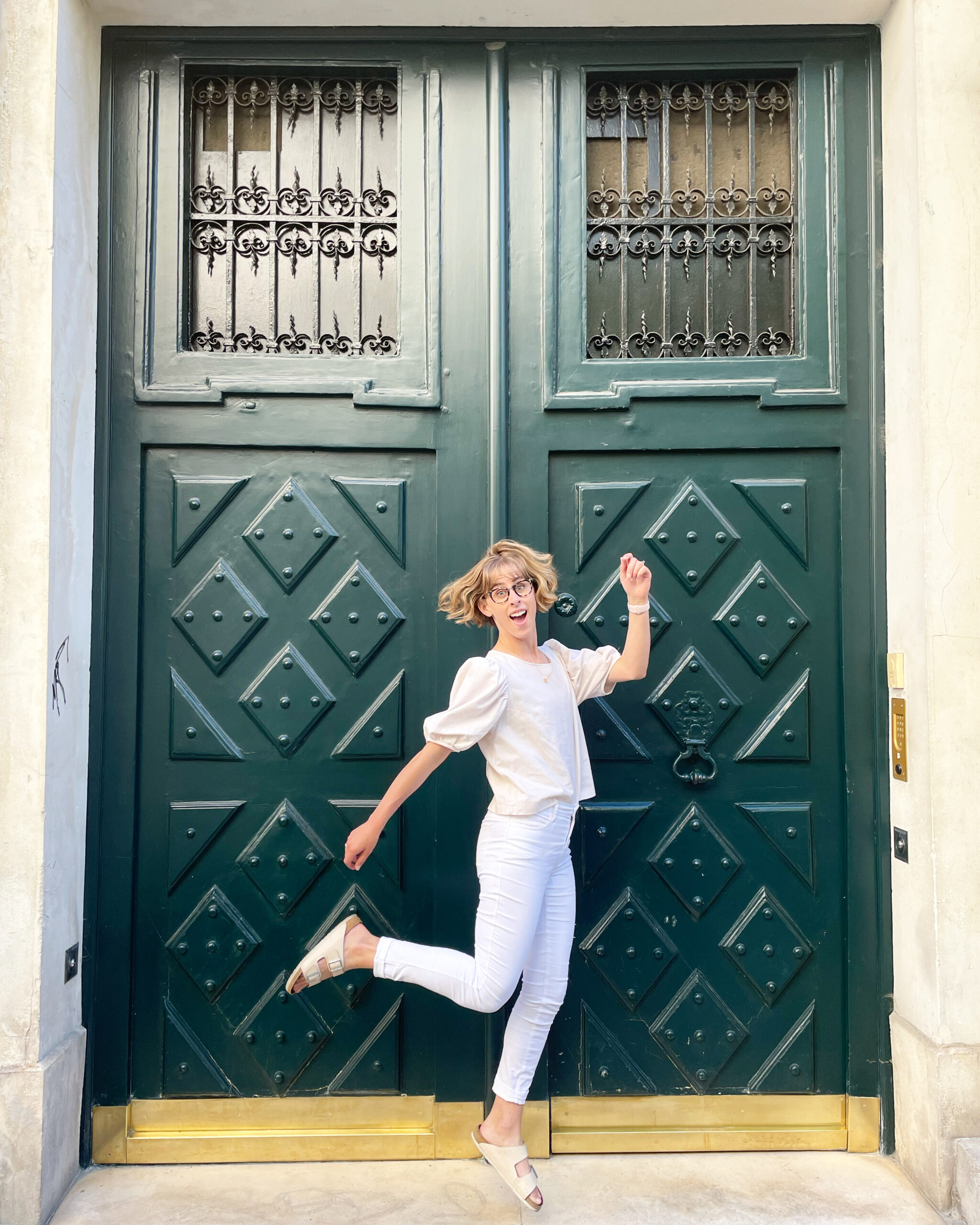 @hann__made, Hannah Hammill, dressed in a white outfit, jumps in front of a pair of large green doors.