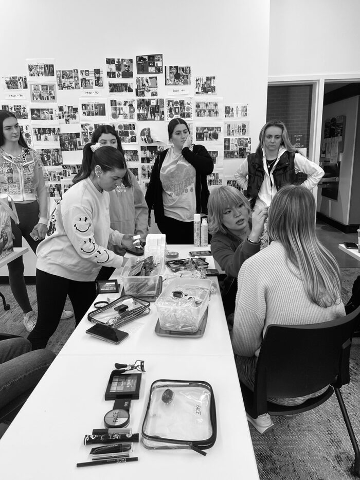 Students watch as woman has makeup applied as part of the SNR36 fashion show prep