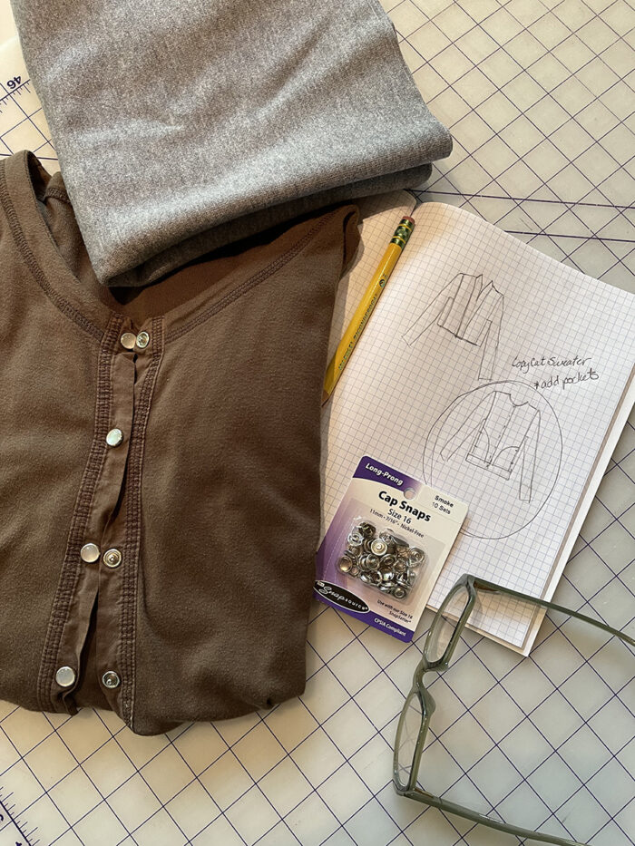 Black cardi with snaps, sketchbook, pattern envelope, gray fabric, and glasses lying on a gridded cutting mat