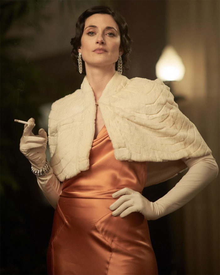 Woman in 1920s ensemble, wearing white stole over silky dress, long white gloves, chandelier earrings and smoking a cigarette