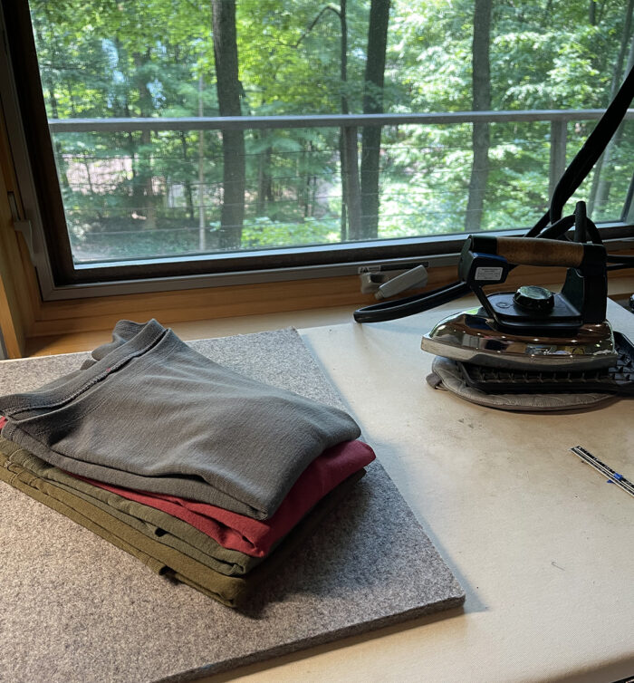 four T-shirts folded and lying on a felt pressing surface with an iron nearby and in front of a window overlooking a wooded area