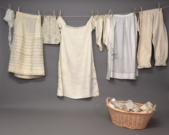 Antique hand-sewn white garments hanging on a washline with a wicker washbasket nearby, part of the Sewn in America exhibit